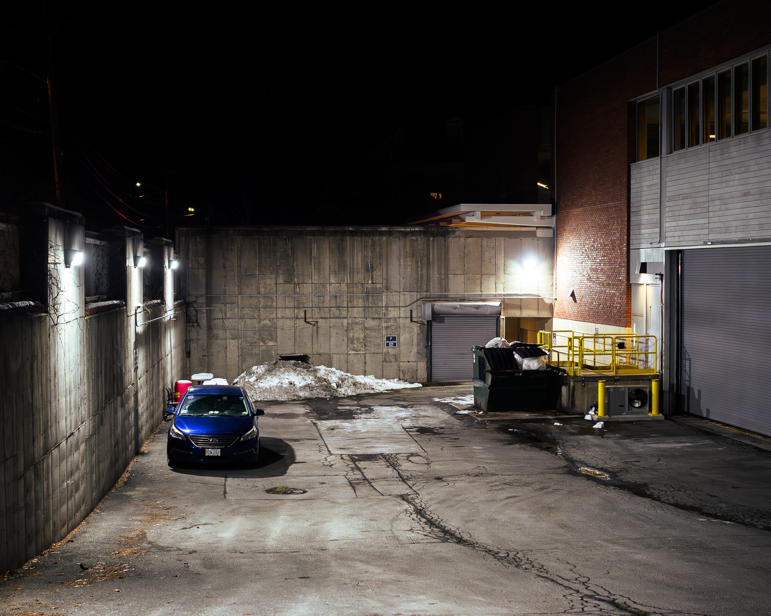 The loading dock after dark: a parked car, a dumpster, concrete retaining walls under floodlight.