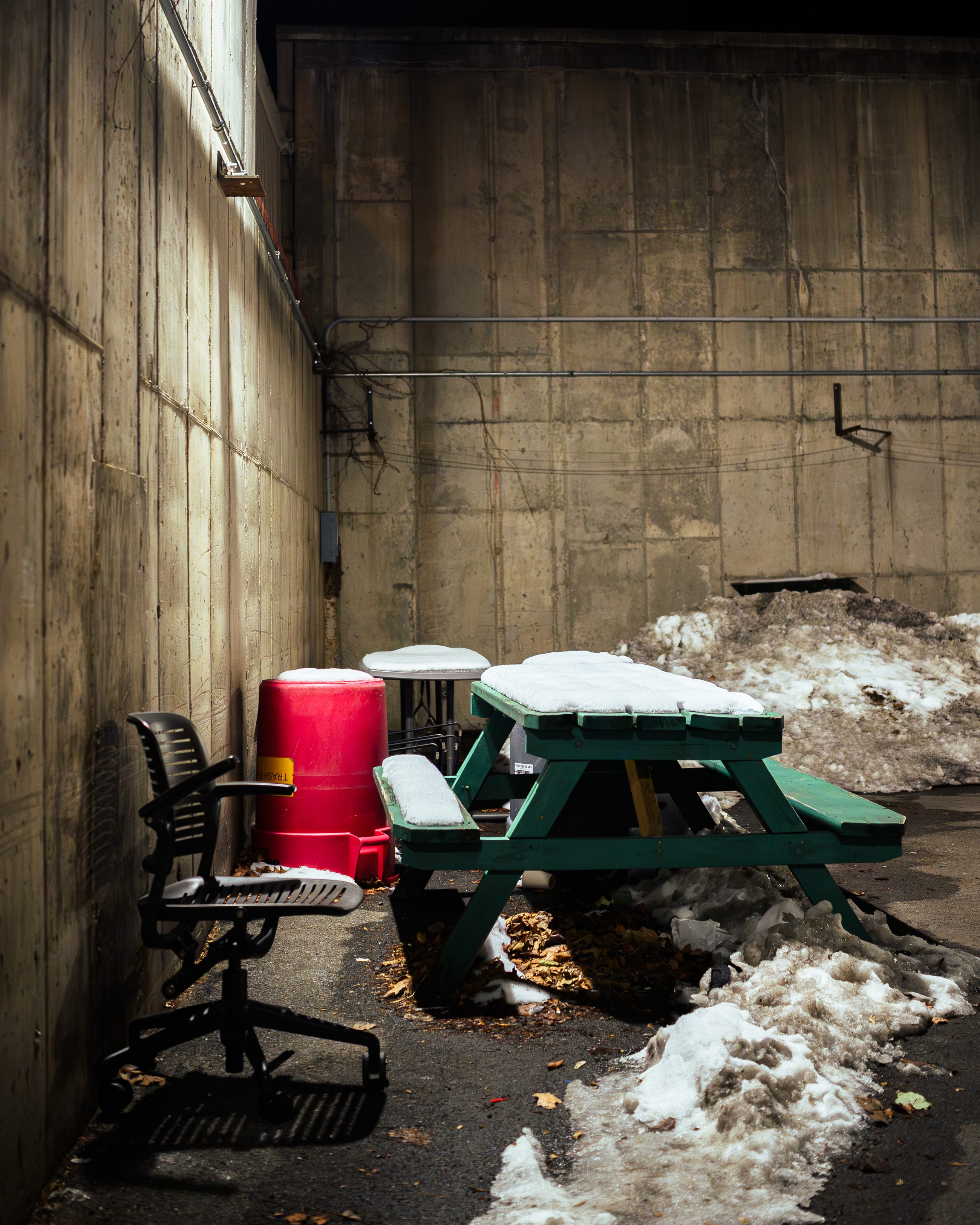 A concrete service yard holding a picnic table, two discarded office chairs and a red bin, snow in the corners.