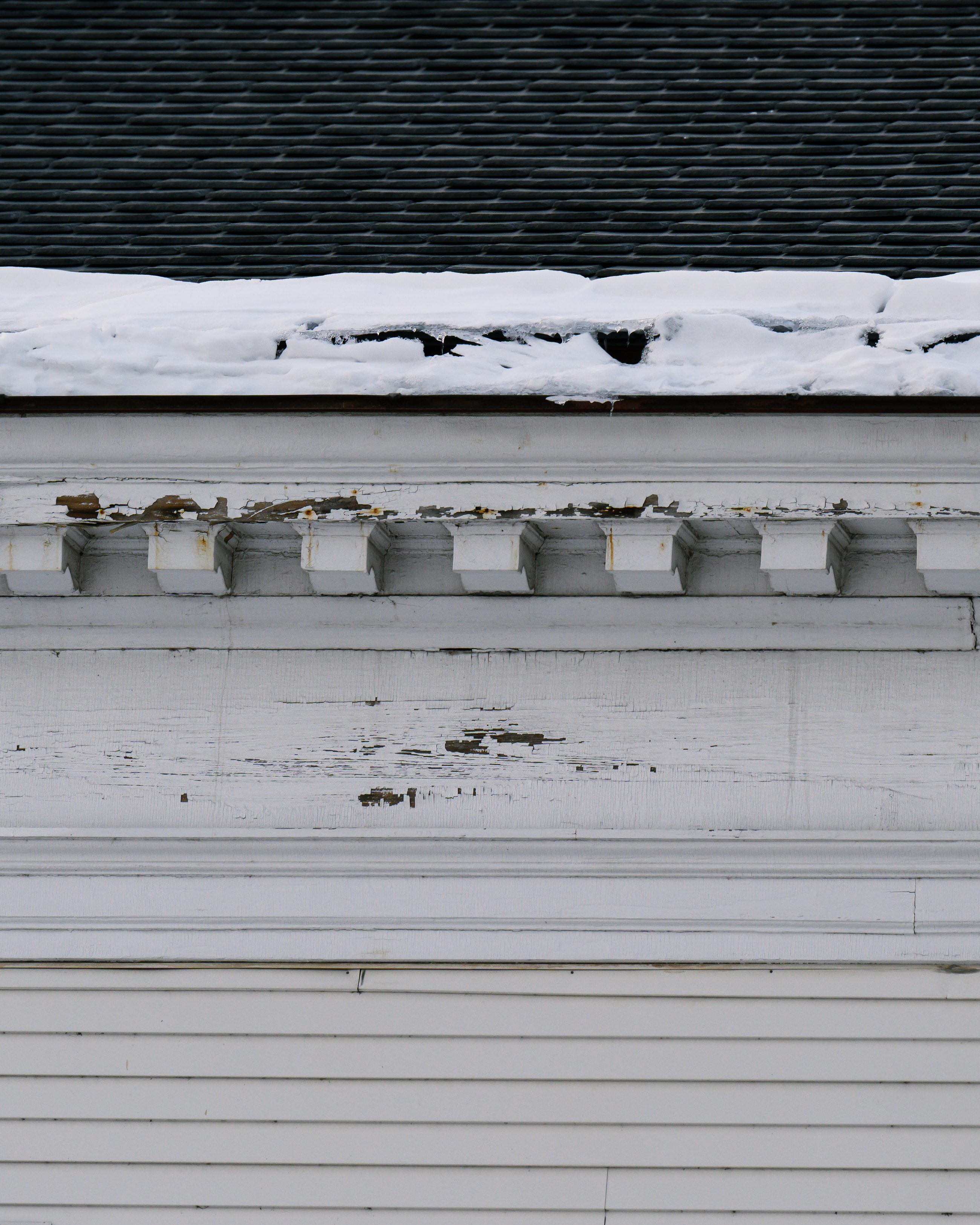 The church's clapboard flank in flat light, snow banked on the roof edge, paint lifting along the cornice.