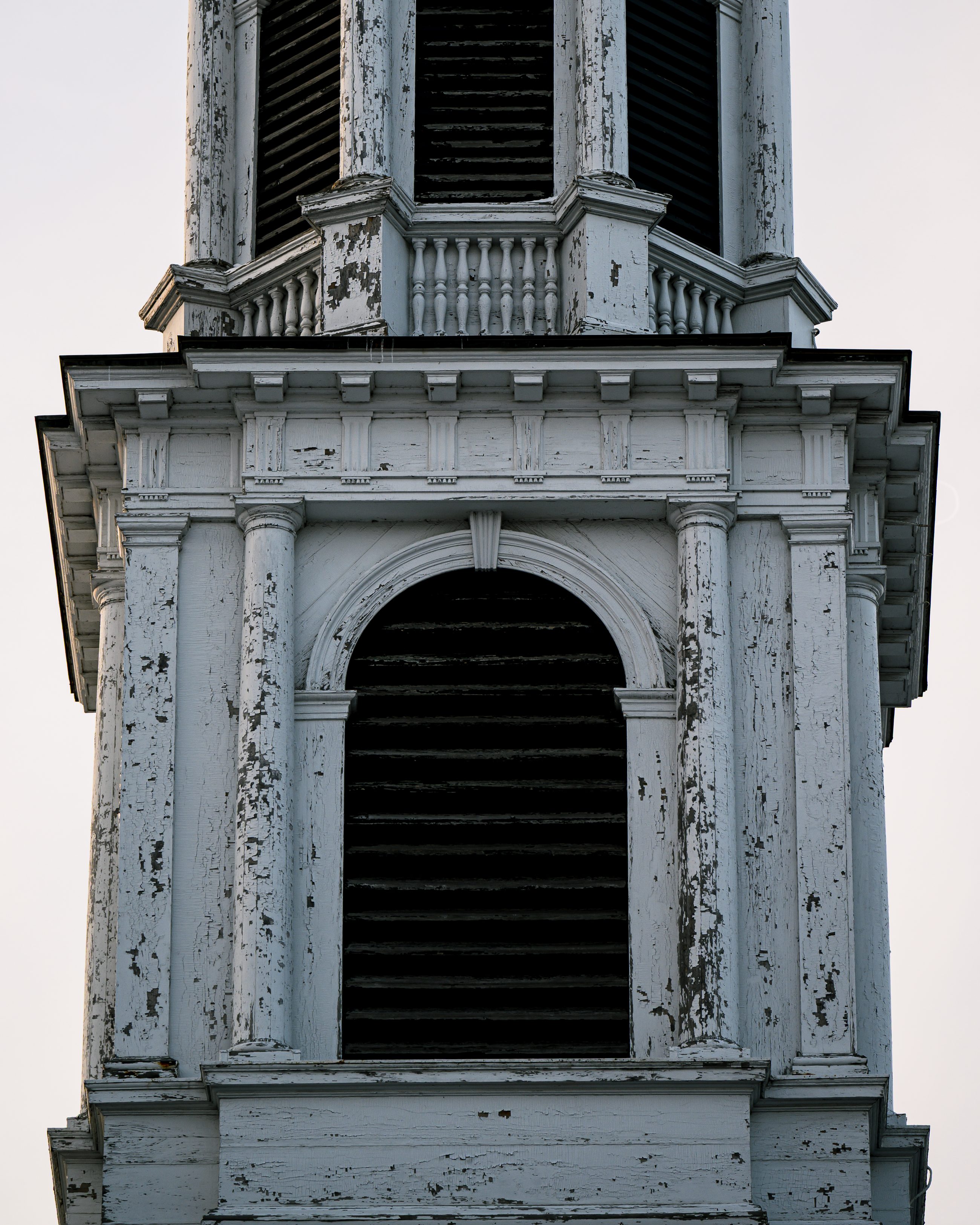The steeple close up: white paint blistered and flaking from the louvred openings and mouldings.