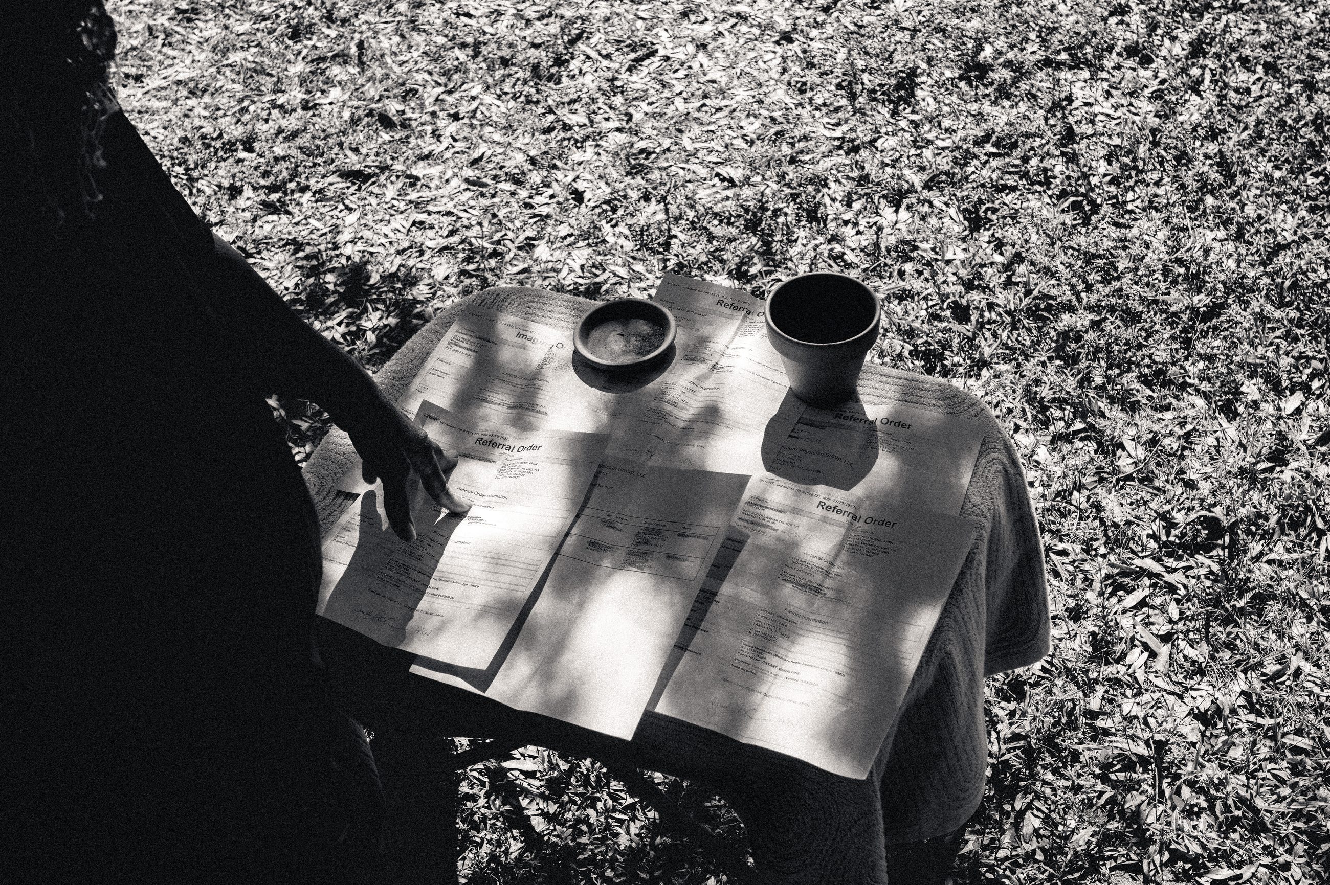 A hand resting on papers spread across an outdoor table in dappled light, two dark vessels beside them.