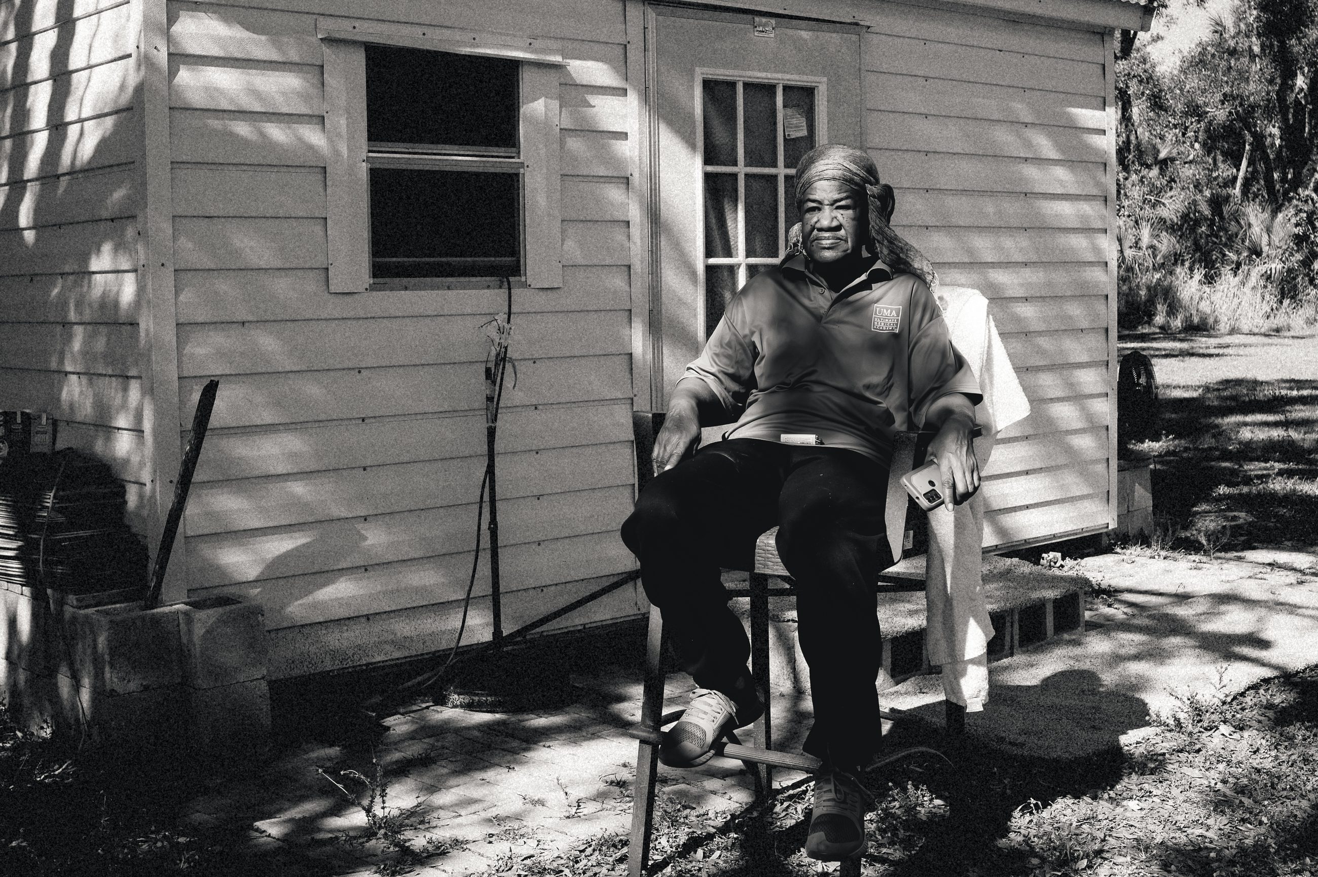 A woman seated on a chair before a small clapboard house, facing the camera directly, a phone in one hand and a towel draped over the chair back; tree shadows fall across the wall behind her.