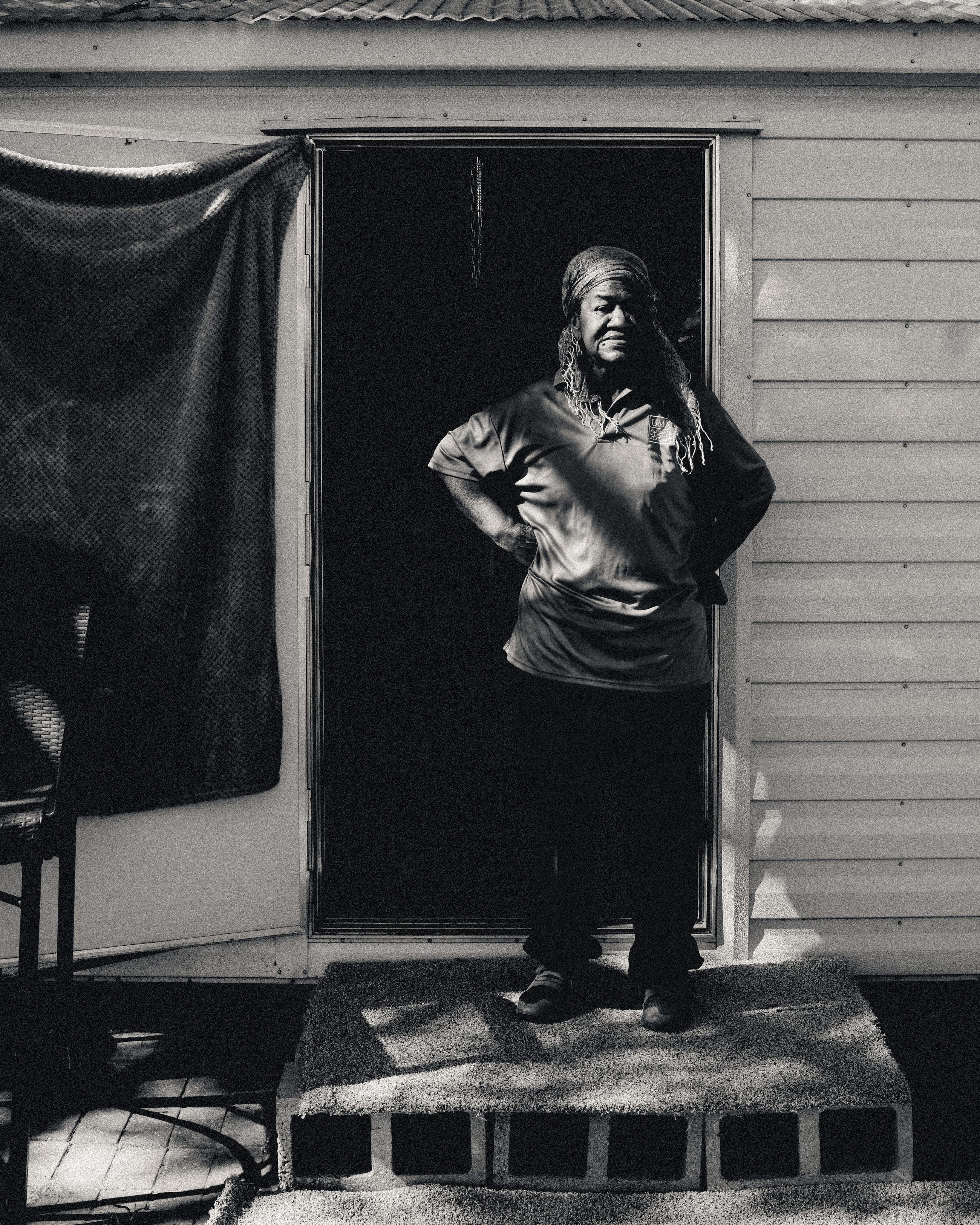 A woman standing in the doorway of her house, one hand raised to her face, dark cloth hung behind her.
