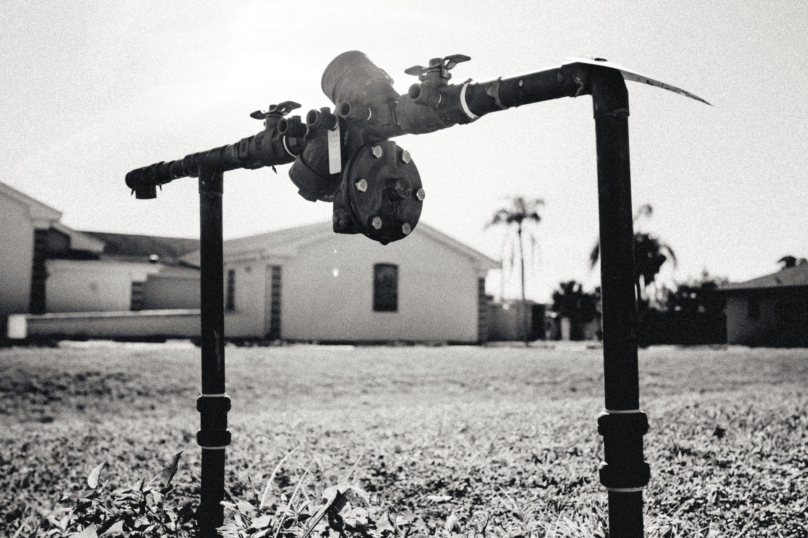 A standpipe and valve assembly rising from a bare yard, single-storey houses and palms behind it.