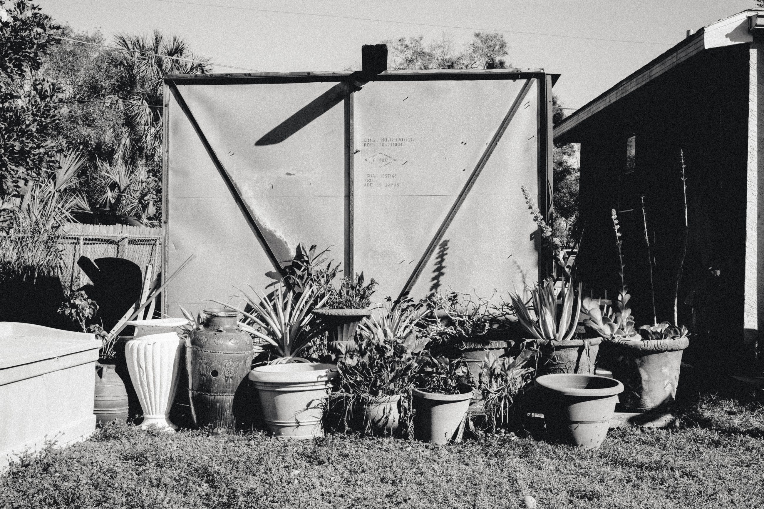 A long row of potted plants along a fence, beneath a tarpaulin frame with a ceiling fan fixed to it.