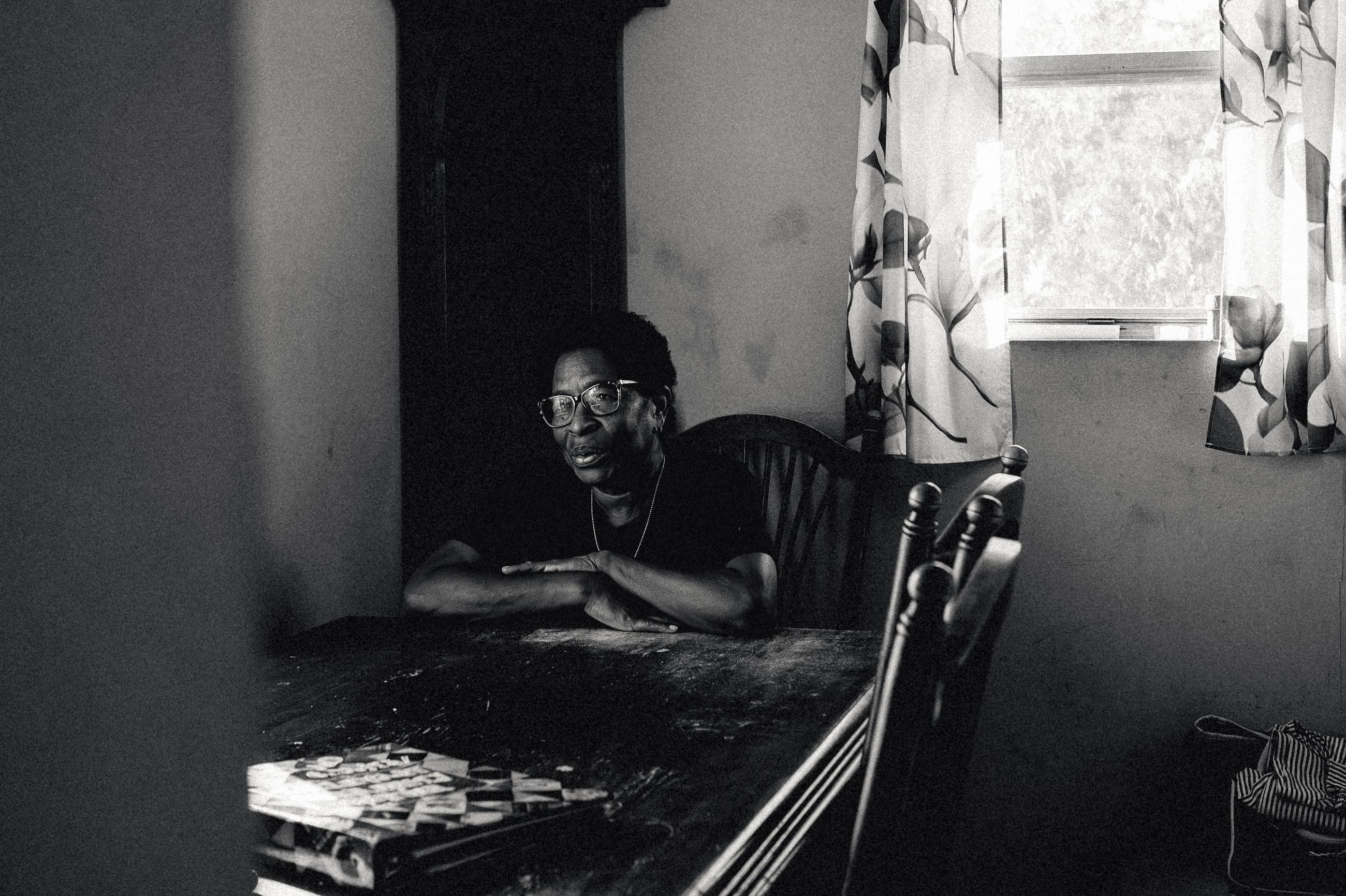 A man seated at a table indoors beside a curtained window, dominoes in front of him.