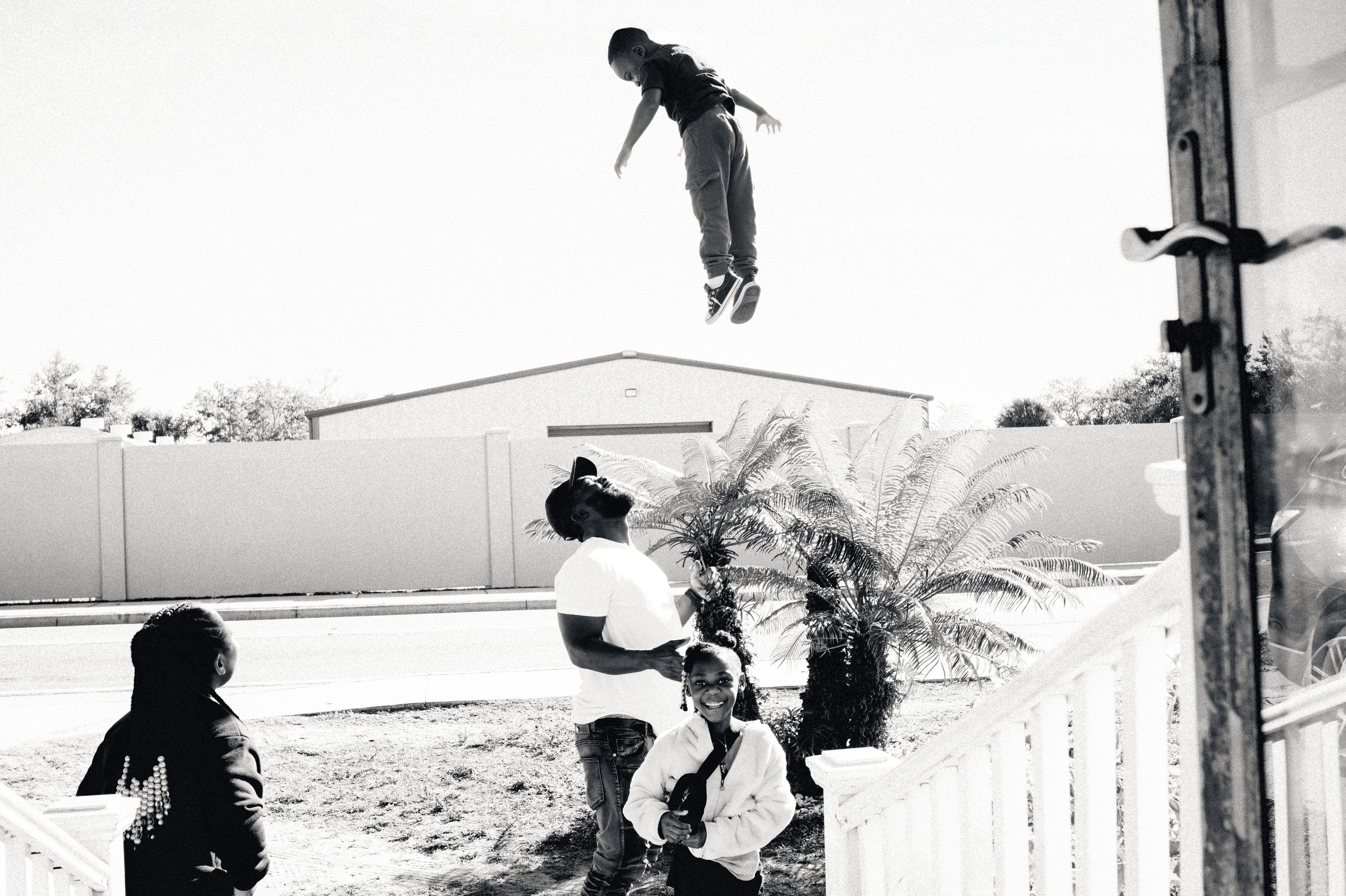 A child in mid-air above a garden fence, other children below, a palm to one side.