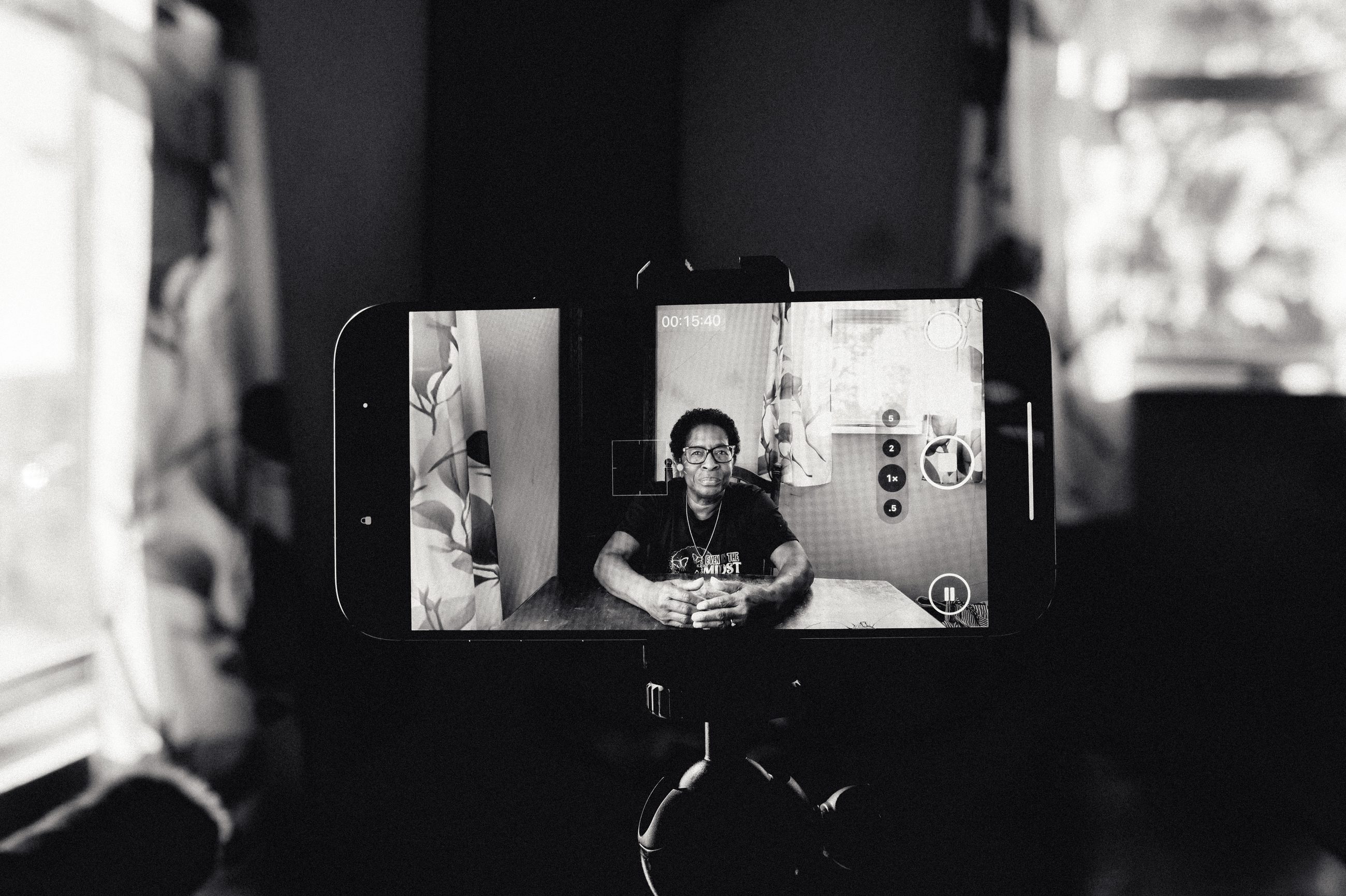 A phone propped on a stand showing a woman on a video call, seated at a table.