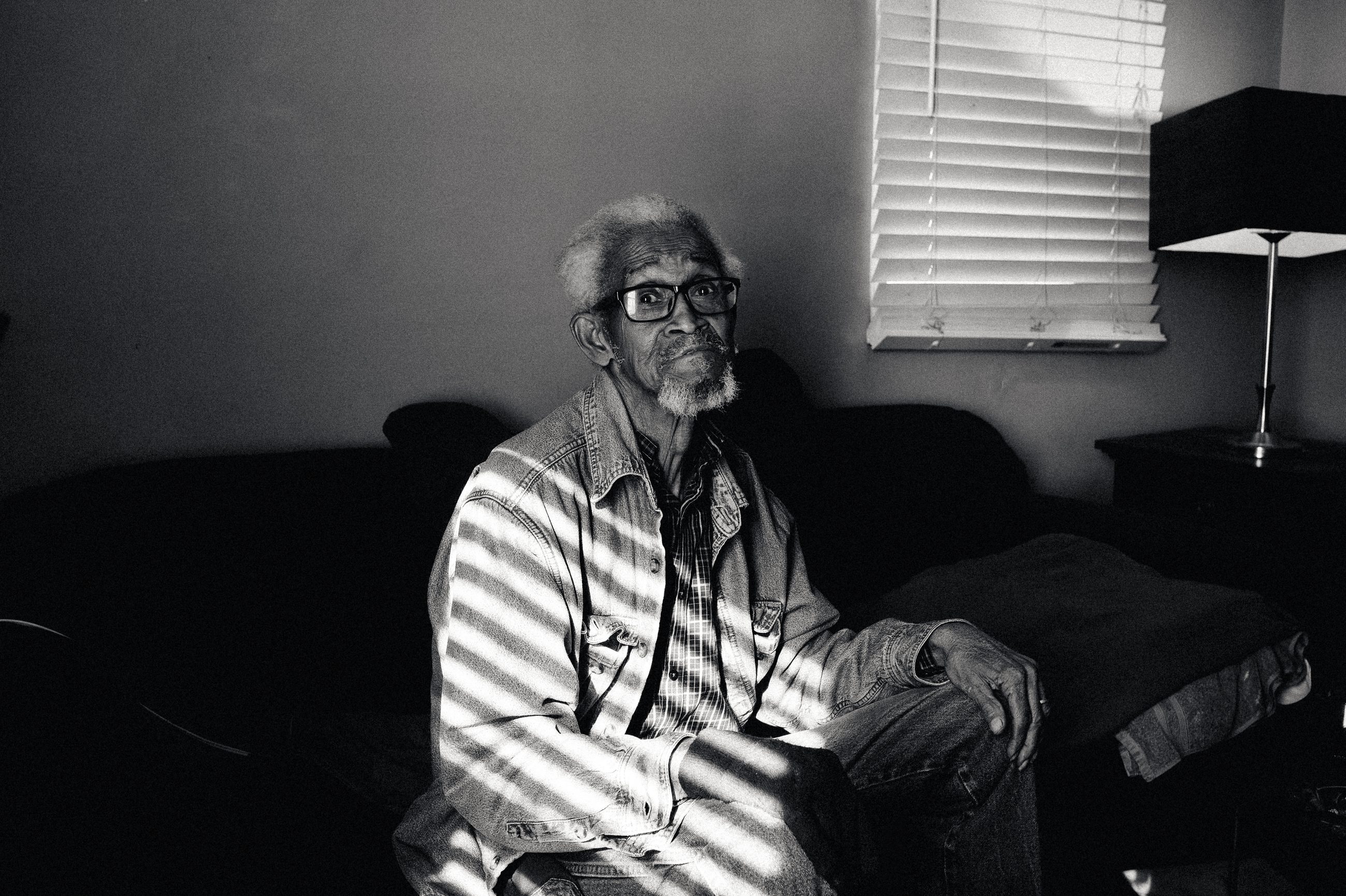 An elderly man in a striped shirt sitting on a couch, light through the blinds behind him.