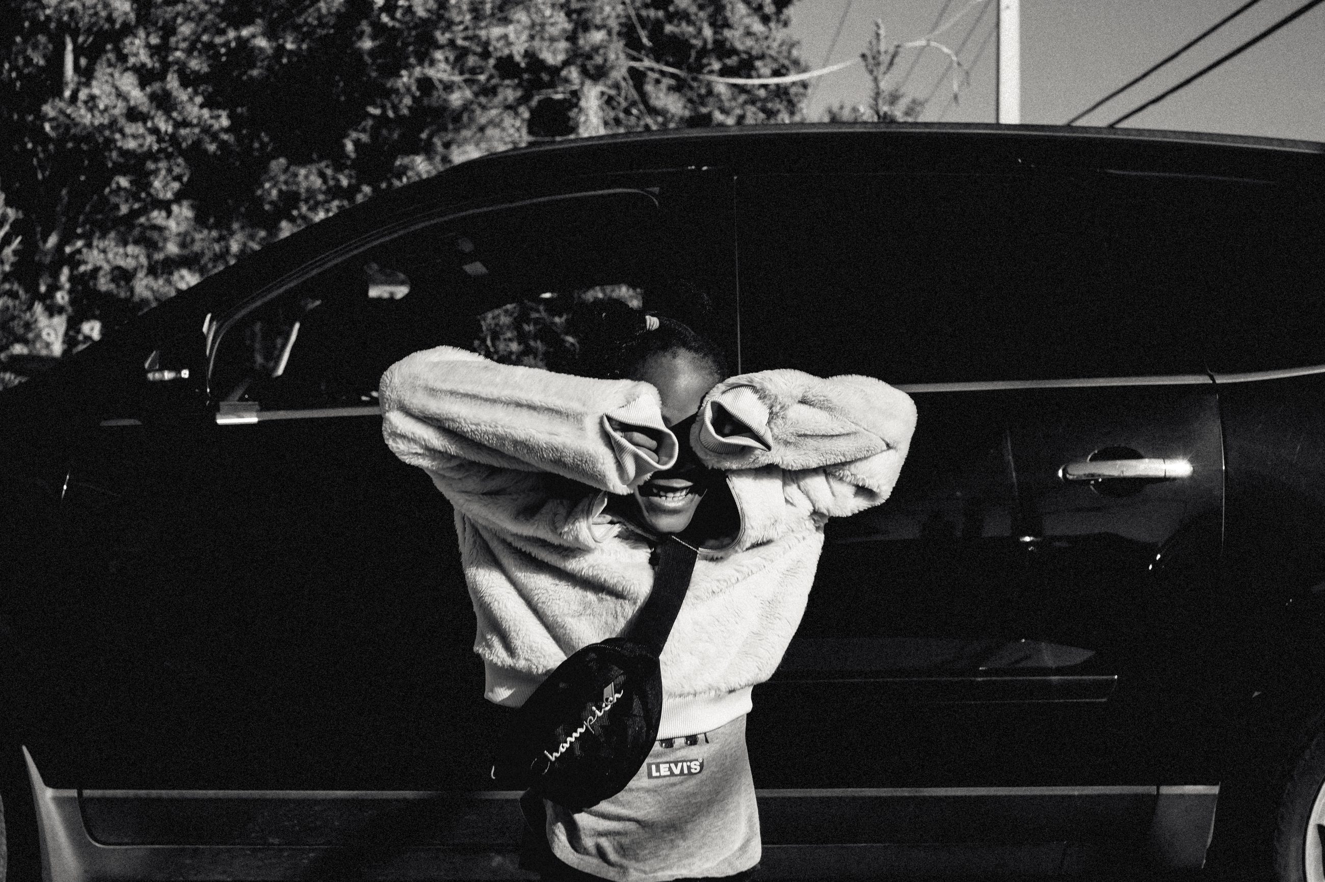 A young person in the doorway of a car, arms raised over their head, holding a cap.
