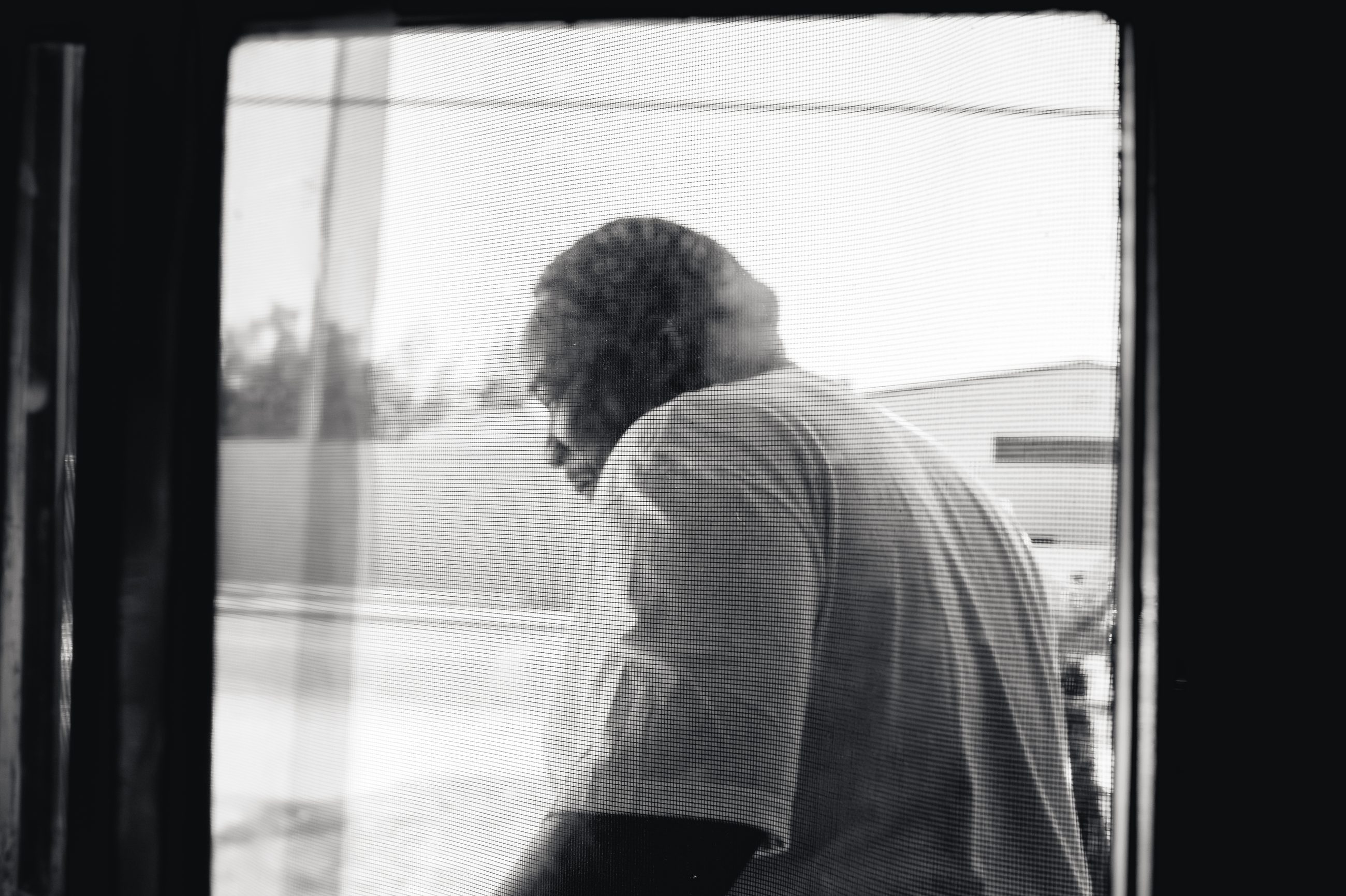 A man bending over his work, seen from outside through a window screen.