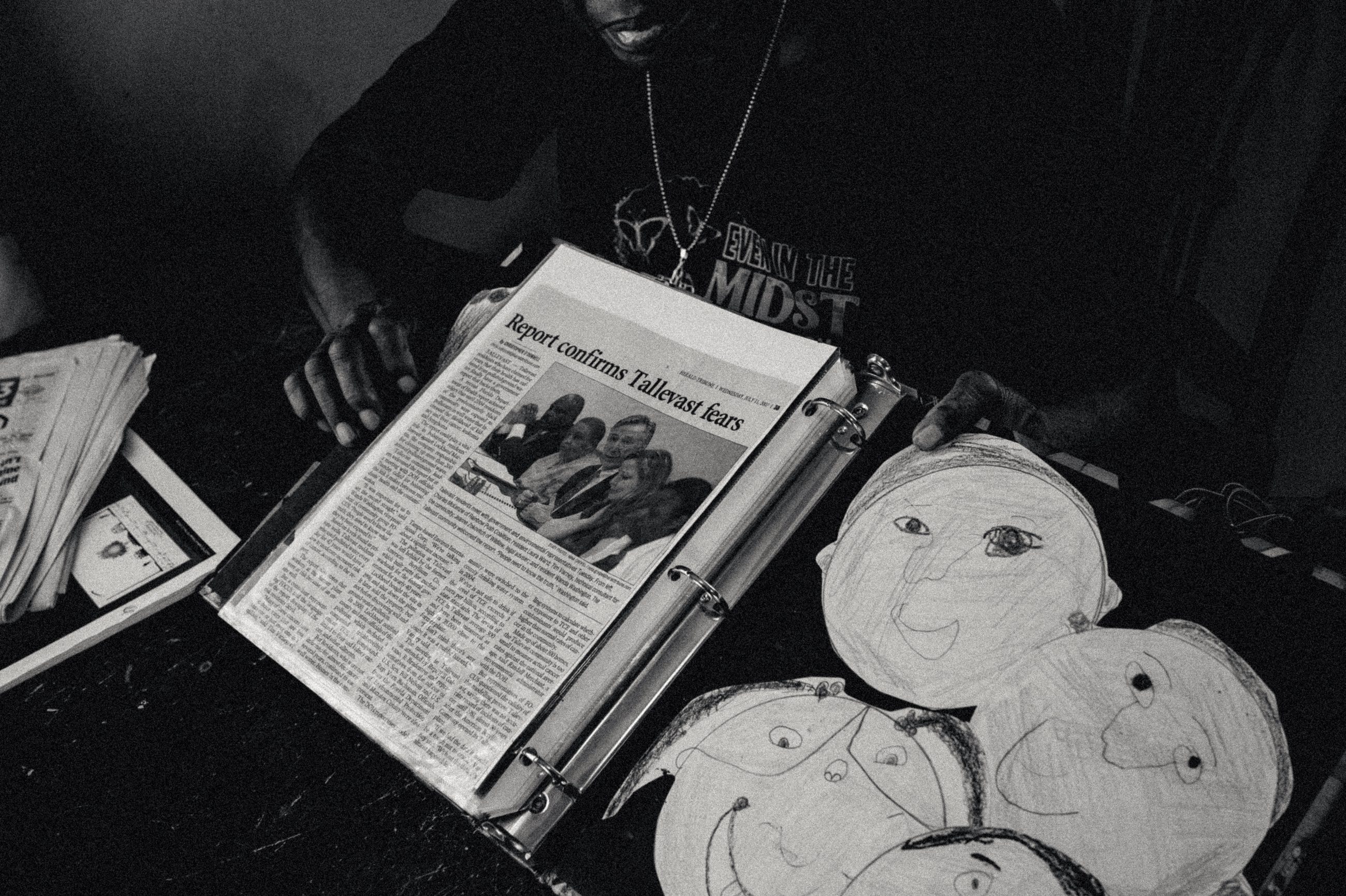 A newspaper report on the Tallevast contamination lying open on a table, beside children's crayon drawings of faces.