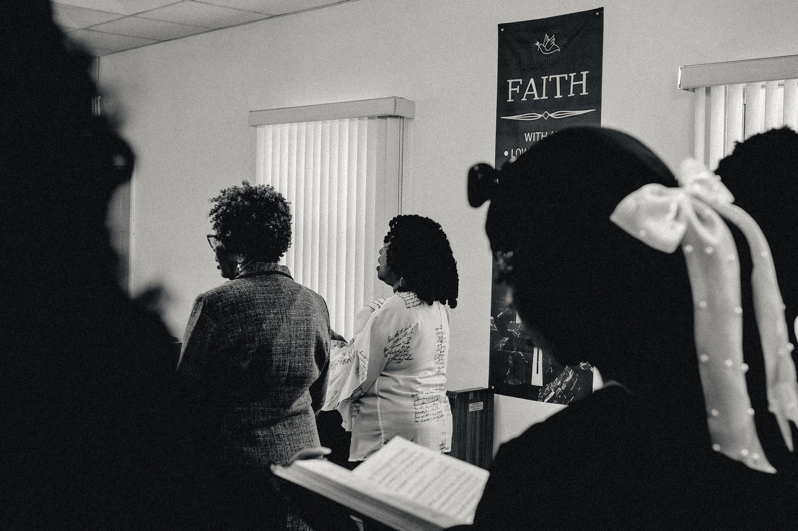 Seen between the silhouetted heads of the congregation: people standing with hymnals, a banner reading Faith.