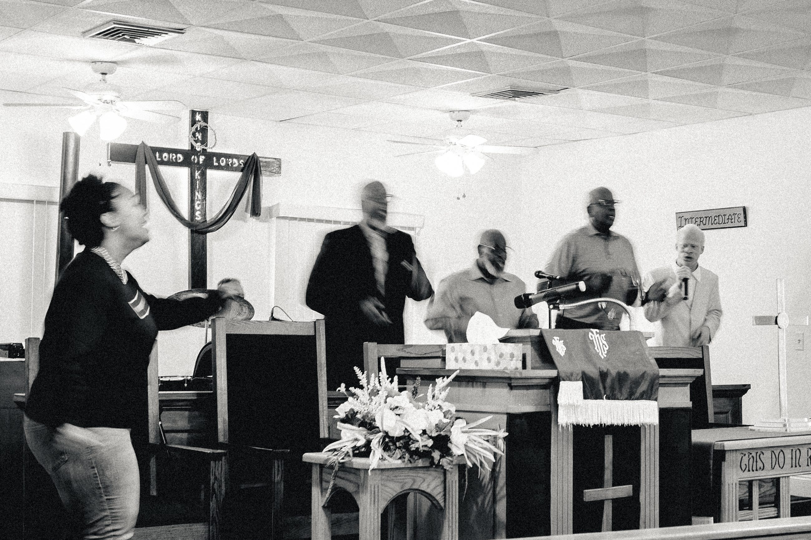 A woman gesturing at the front of the church; men at the pulpit with microphones and instruments.