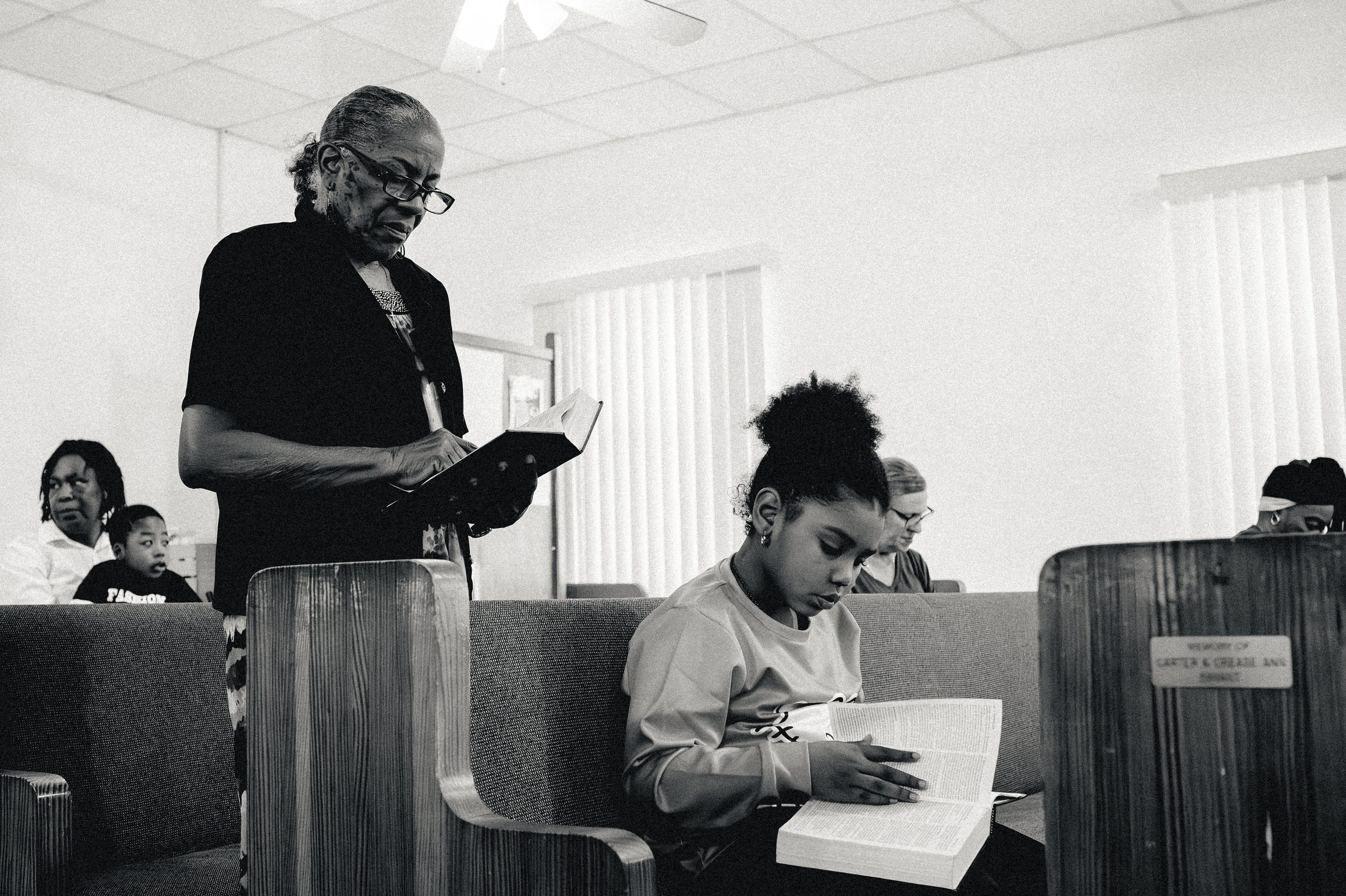 An elder standing to read from a book. A young woman sits beside him, reading from her own.