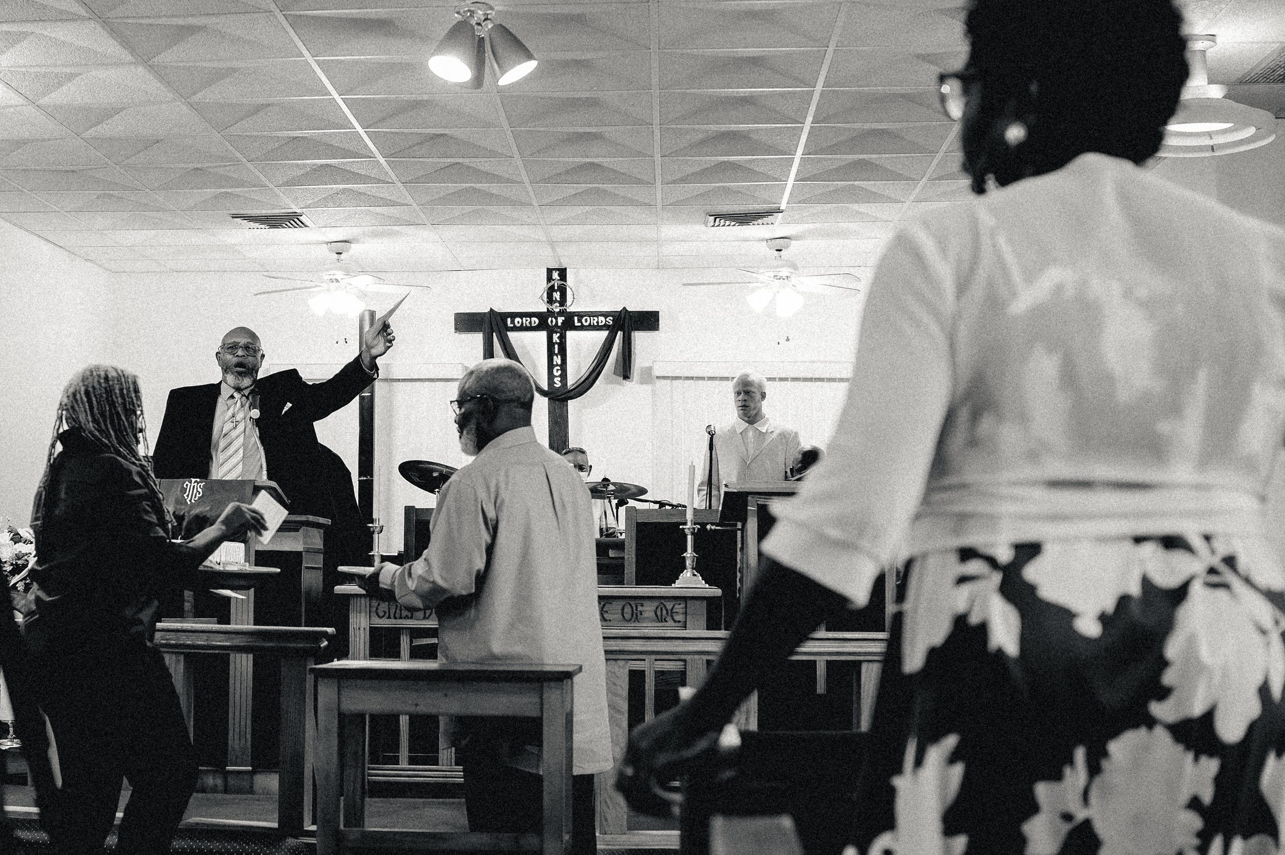 A figure passing close to the camera, blurred; beyond her a man at the front with his hand raised.