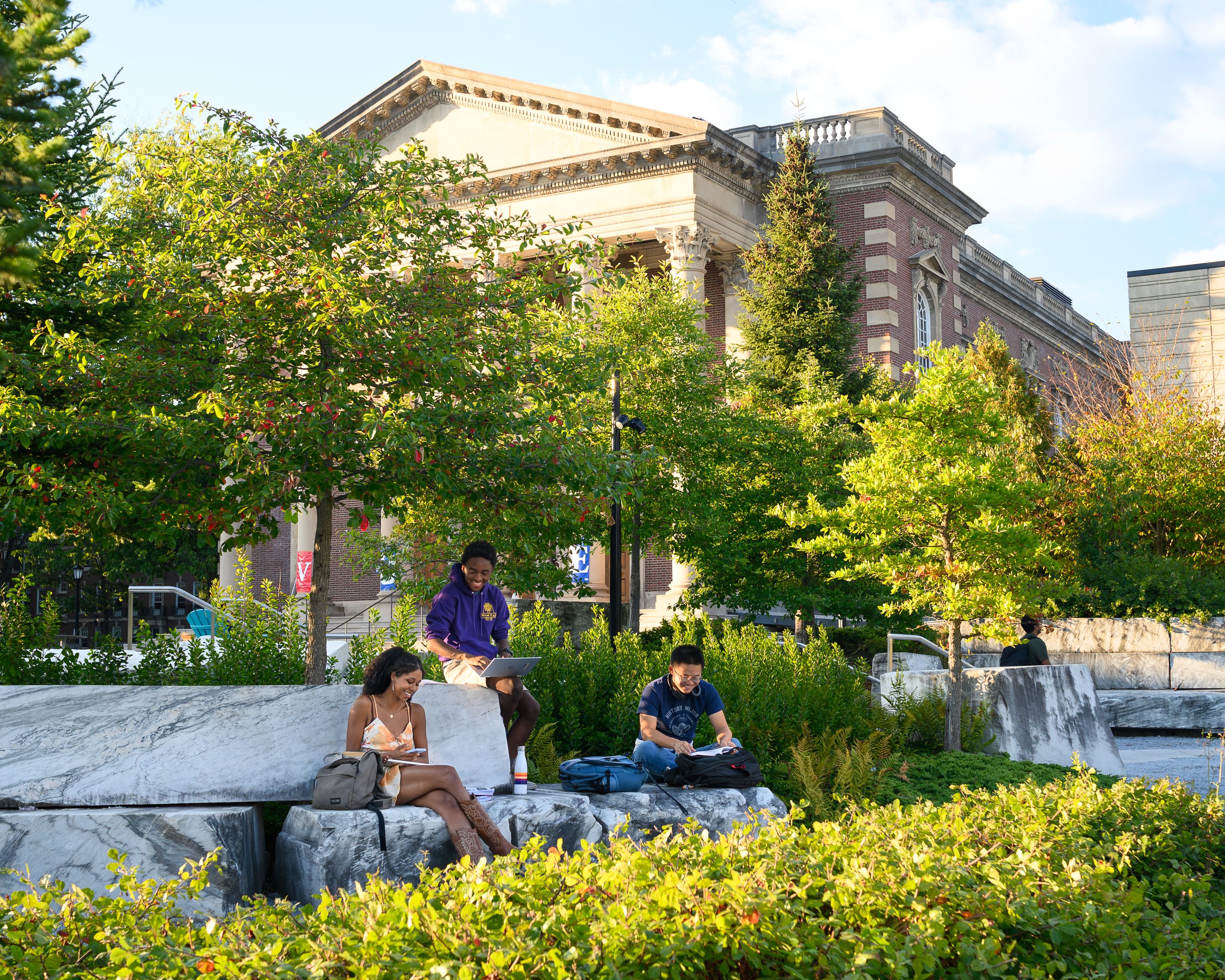 Students sitting and talking on low stone blocks among planted greenery, a neoclassical facade behind them.