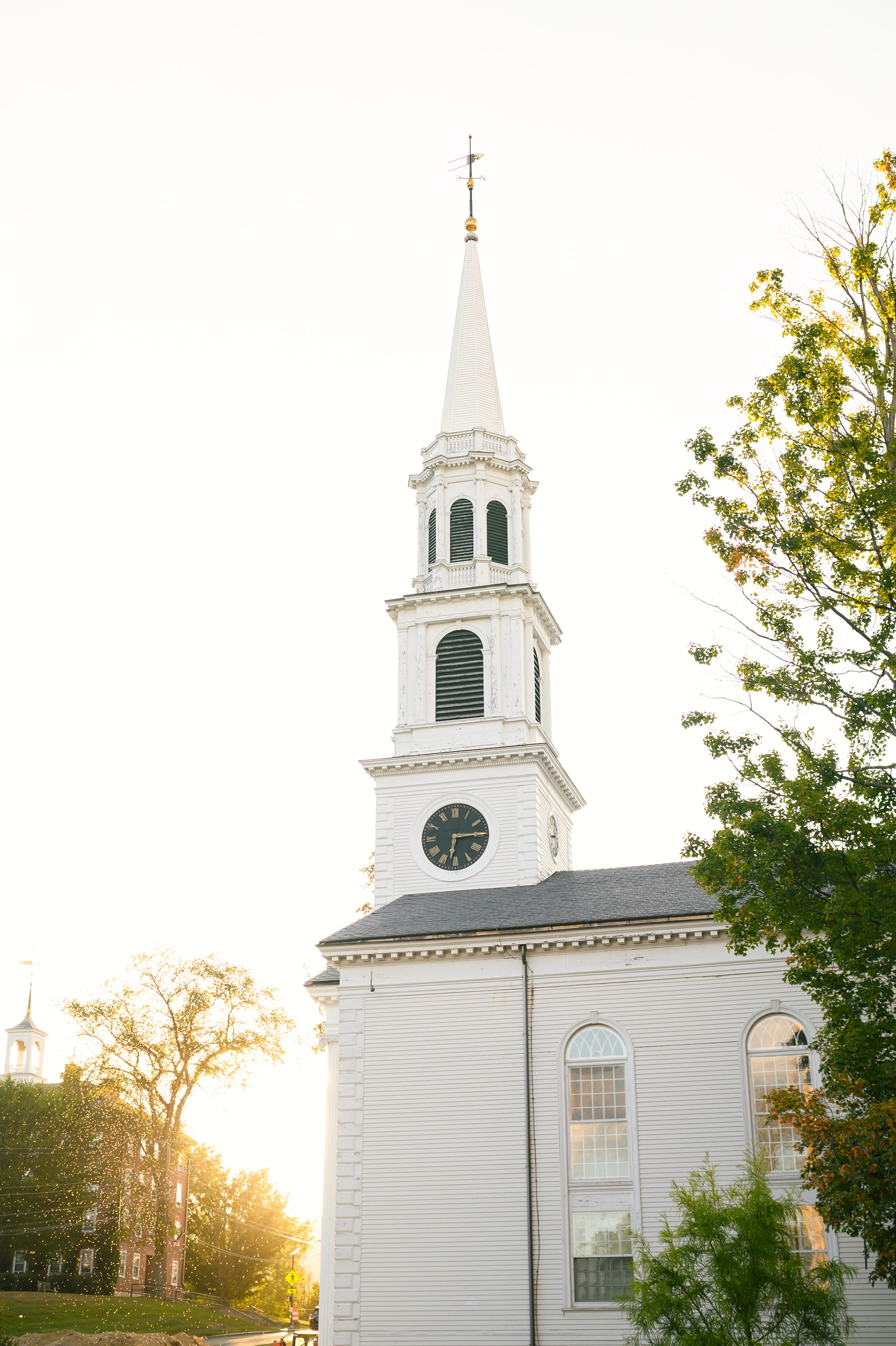 The white church steeple against a warm sky at the end of the day, treetops below it.