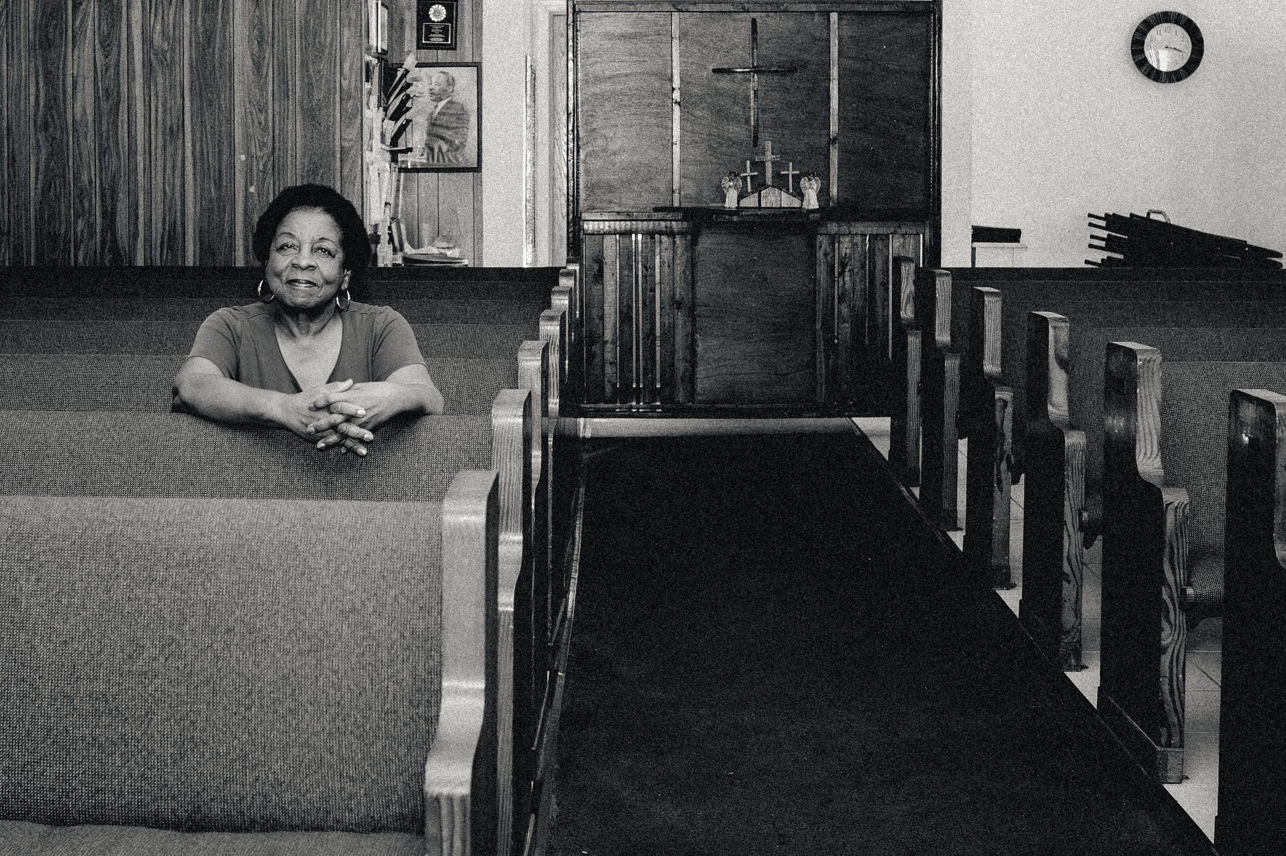 A woman leaning on the back of a pew in the empty church, the aisle running away behind her.