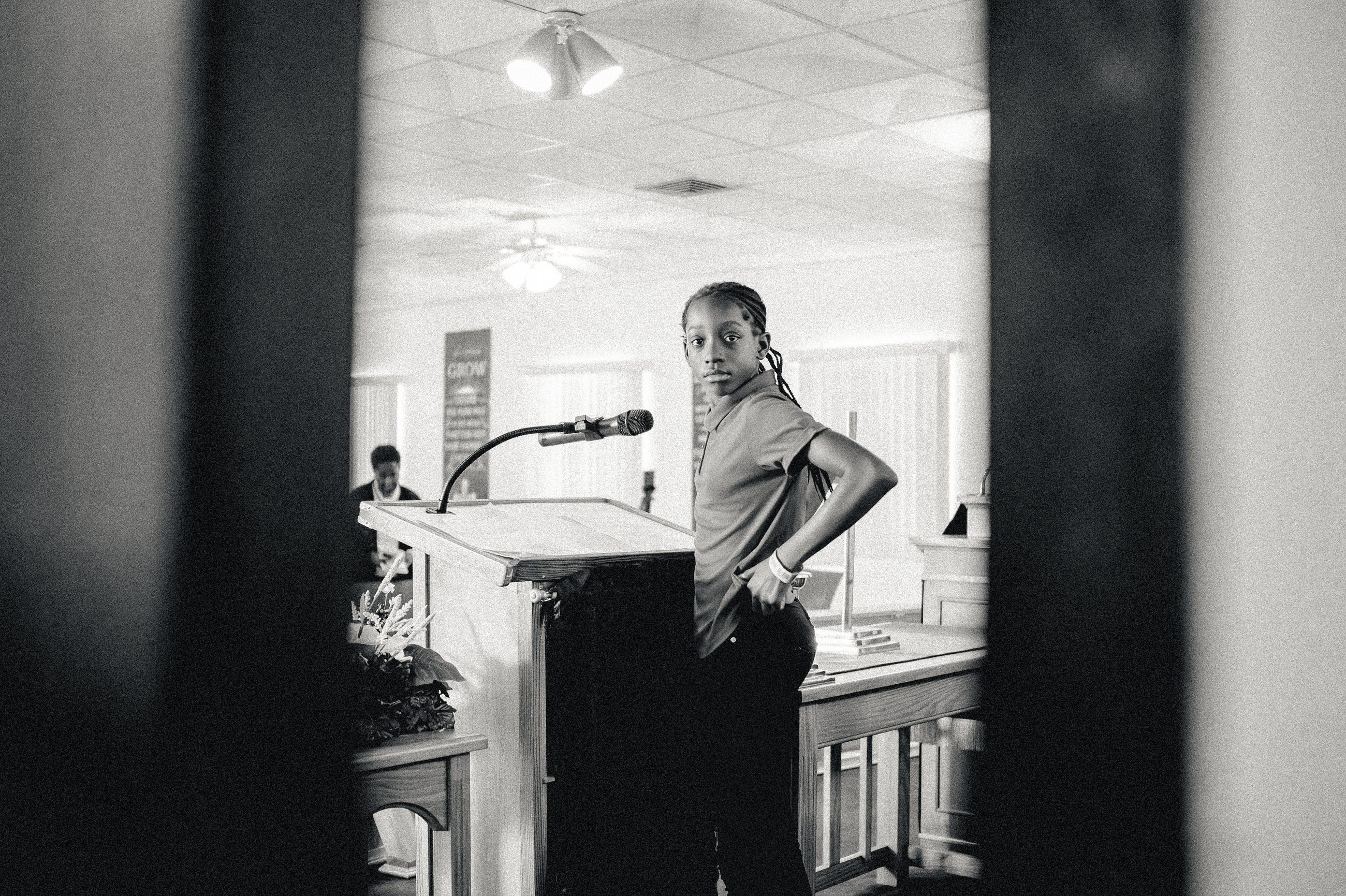 A girl standing at the lectern, framed by a dark doorway.