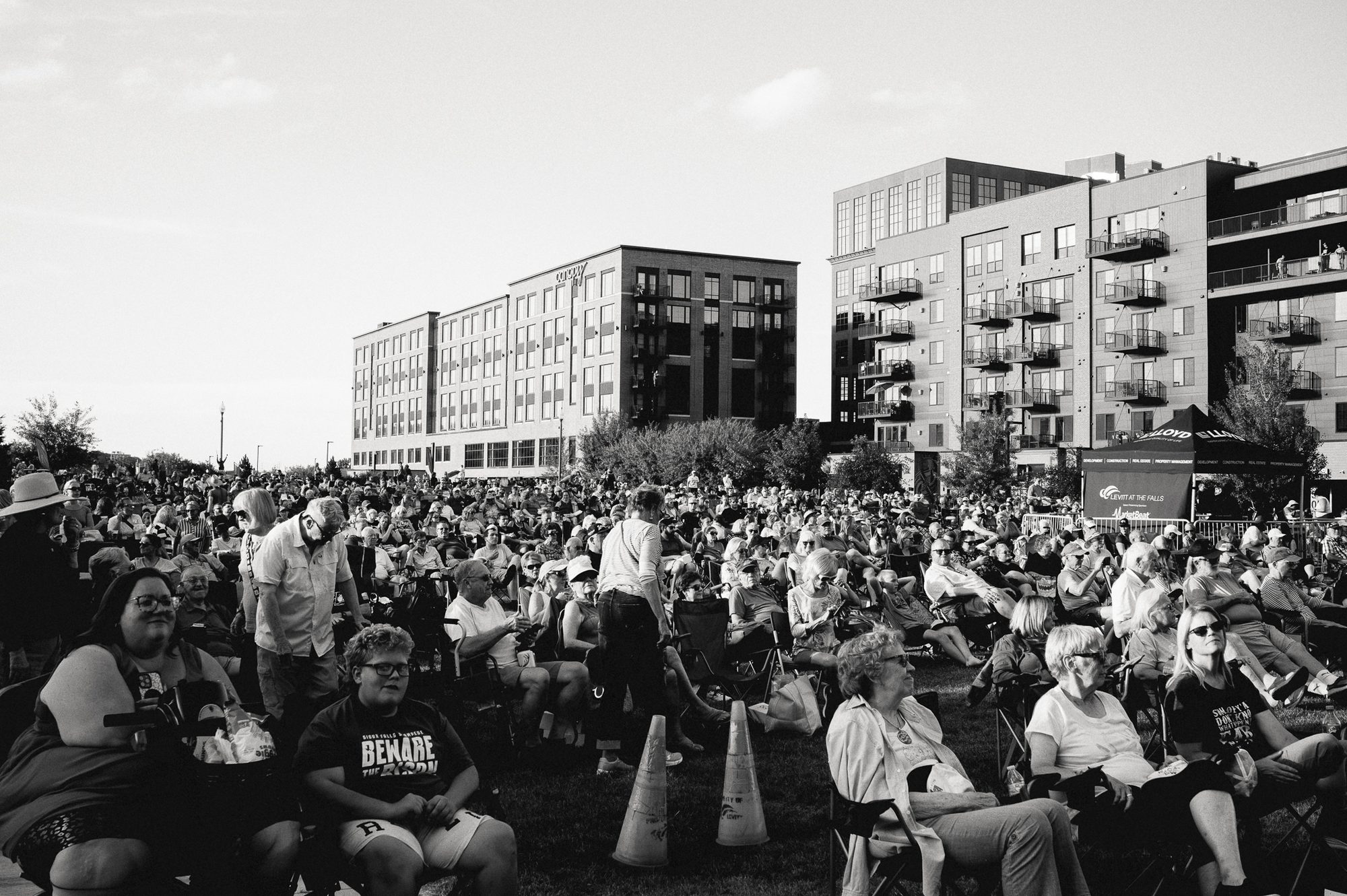 A crowd seated on lawn chairs across a field at an outdoor concert, all facing the same direction, apartment blocks rising behind them.