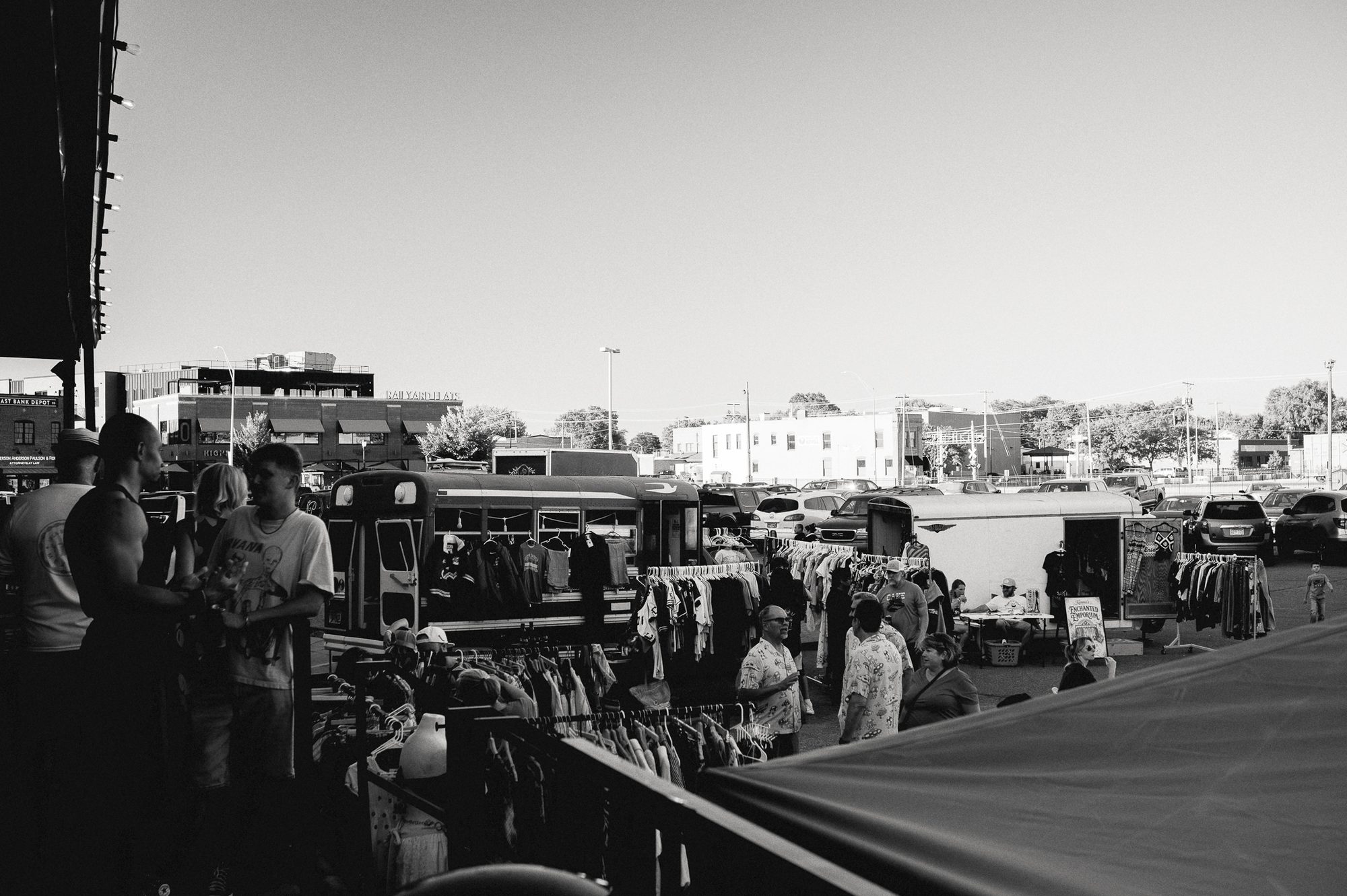 A street market seen over a parked car: stalls of clothing, people browsing, traffic beyond.