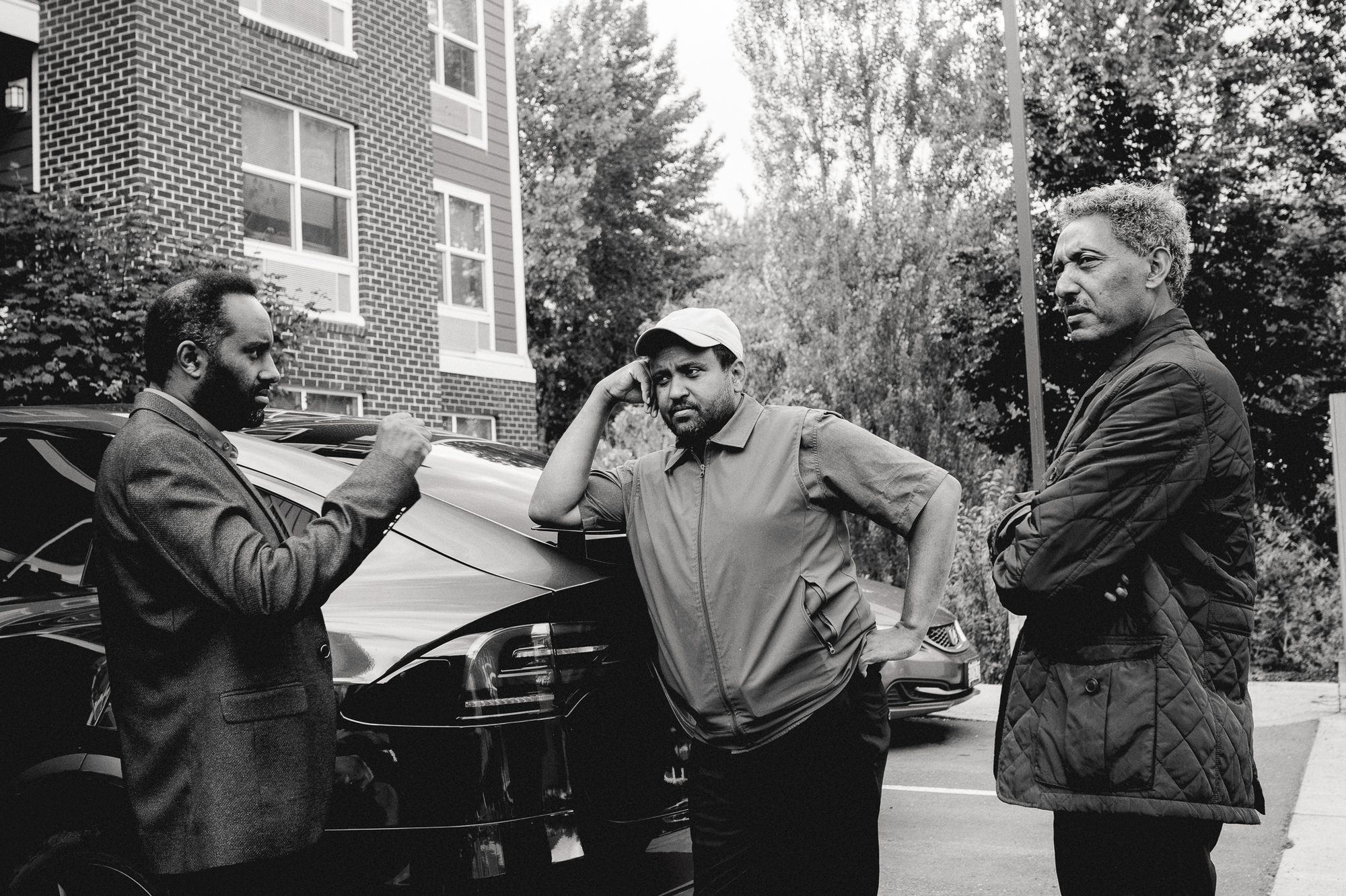Three men talking on the pavement beside a parked car, one leaning on the roof.