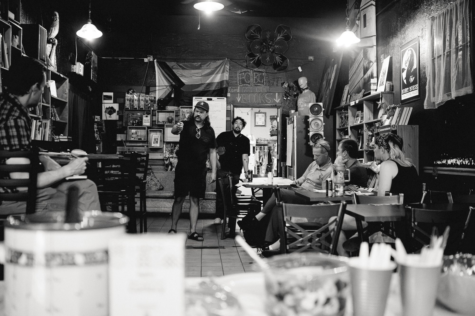 A bar interior in the late afternoon, people at tables under hanging lamps, glasses and a jar in the foreground.