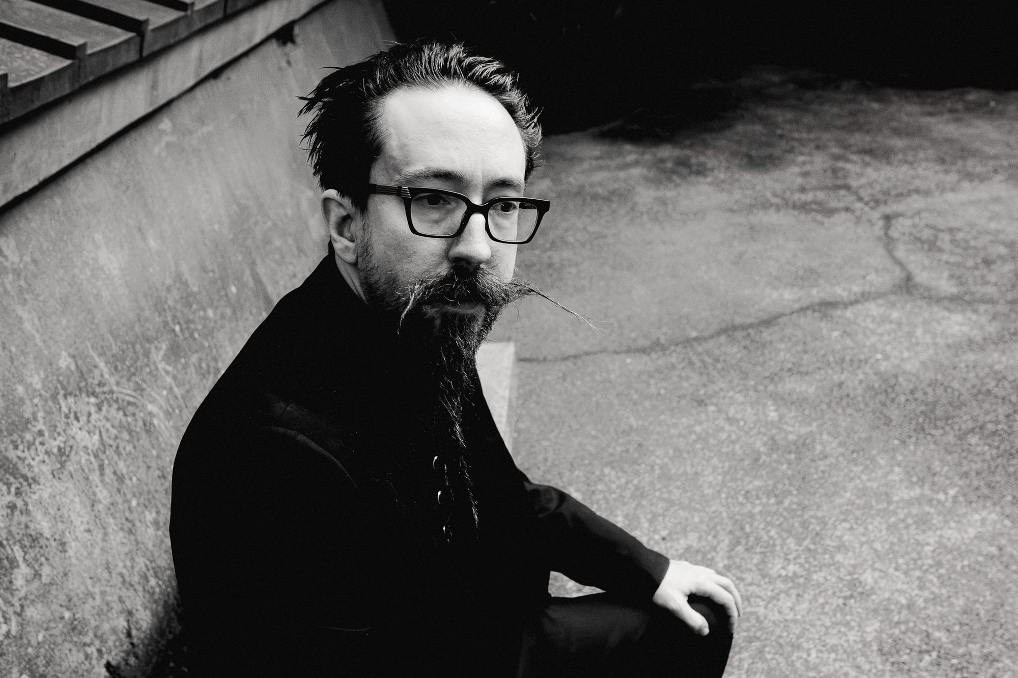 A man in glasses against a concrete wall, turned slightly toward the camera.
