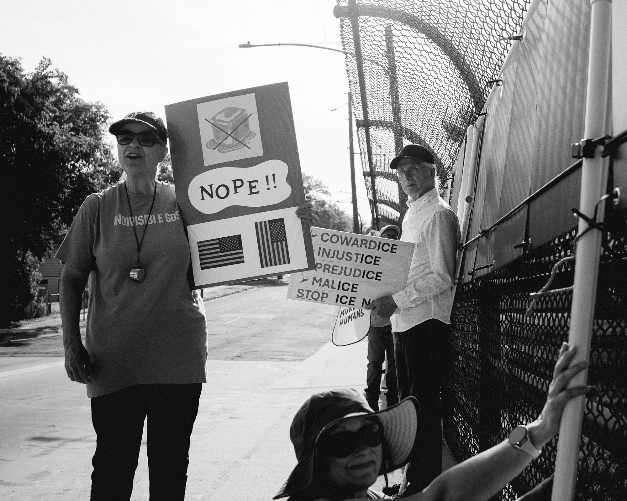 People standing at a chain-link fence on an overpass, holding a hand-lettered sign reading NOPE and, below it, COWARDICE INJUSTICE PREJUDICE MALICE — STOP ICE.